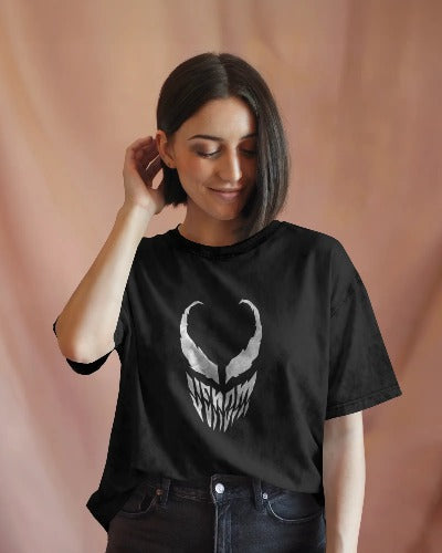 Front view of a female model wearing a black acid wash t-shirt with a white graphic of the Venom mask/face on the chest. The model is touching her hair, posing in a soft-lit, relaxed, and casual style.