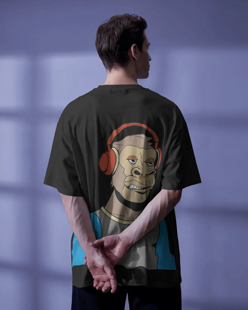 Back view of a male model with dark hair, standing with hands folded behind his back, wearing a black oversized t-shirt. The shirt features a large, stylized graphic of an ape-like character wearing red headphones, looking over his shoulder. The background is a solid purple.