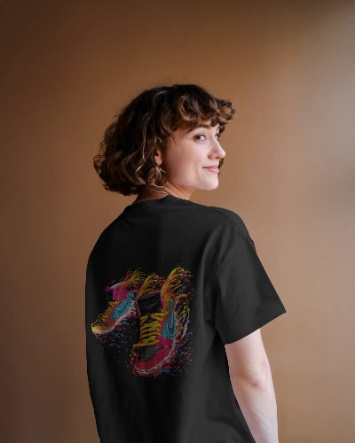 Rear view of a smiling woman with short, curly hair modeling a black t-shirt against a soft brown backdrop. The shirt's back is detailed with a vibrant, paint-splattered graphic of overlapping sneakers.
