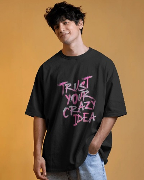 Front view of a male model with dark, messy hair wearing a black oversized t-shirt and blue denim jeans. The shirt's front features the phrase "TRUST YOUR CRAZY IDEA" in bright pink script. The background is a solid orange-yellow color.