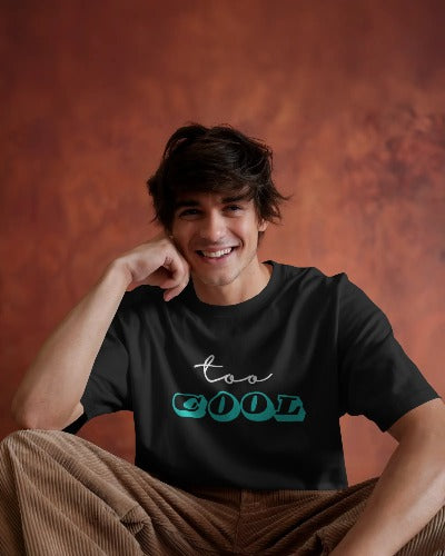 Front view of a smiling male model with dark hair, sitting and resting his arm on his knee. He is wearing a black t-shirt with the phrase "TOO COOL" in light blue and green script on the chest. The background is an earthy brown color.