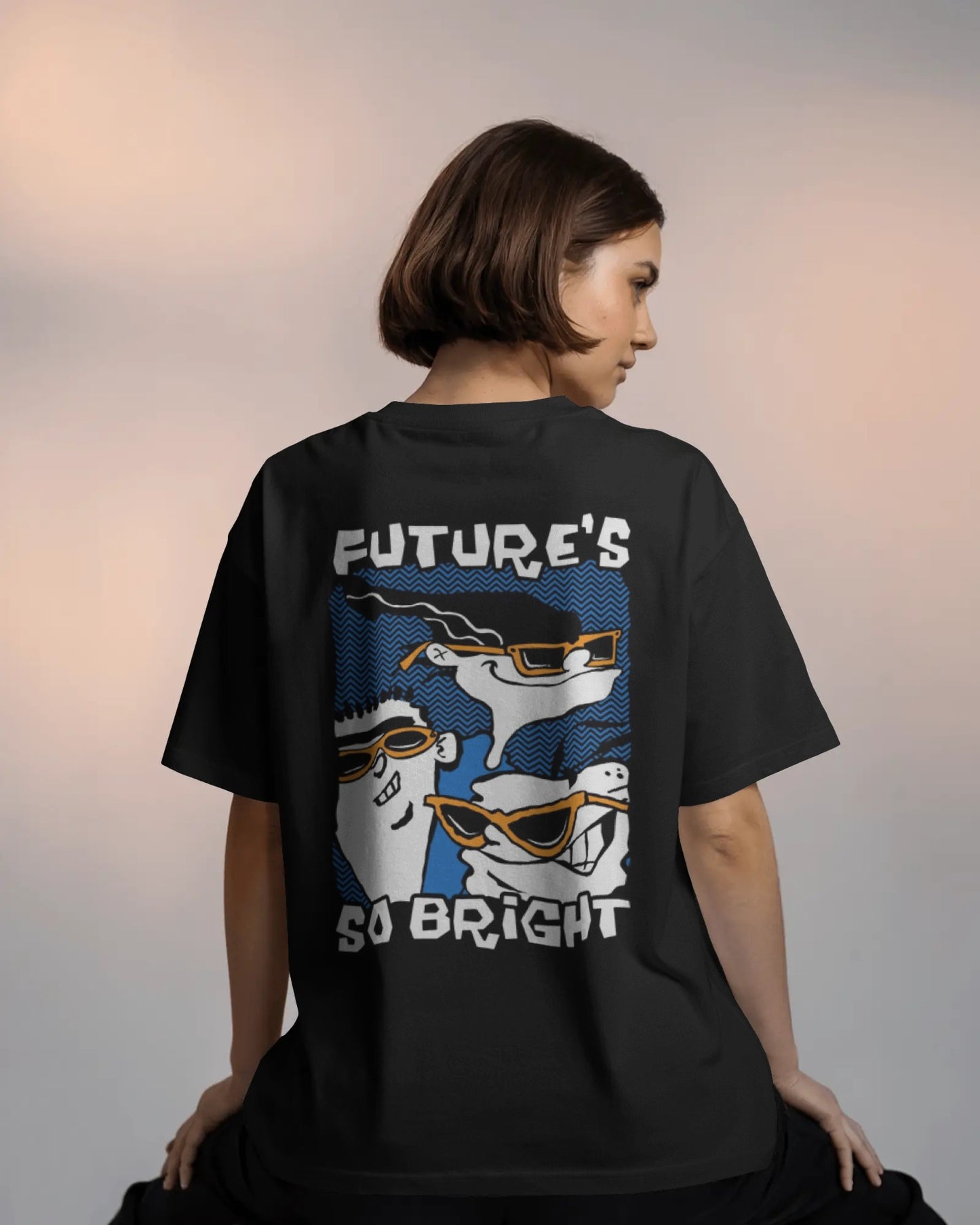 Future So Bright Gotta Wear Shades Oversized T-Shirt - Back view of a model with short dark hair wearing a loose-fitting black t-shirt. The back of the shirt features a large graphic of two cartoon characters wearing sunglasses and caps, with the text "FUTURE'S SO BRIGHT" above them in a white and blue retro style.