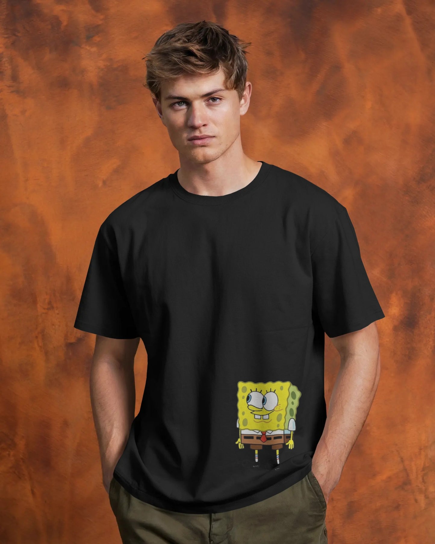 Front view of a male model wearing a black t-shirt and olive pants against a textured orange background. A small graphic of a happy SpongeBob SquarePants peeking out from a mock pocket is printed on the lower front of the shirt.