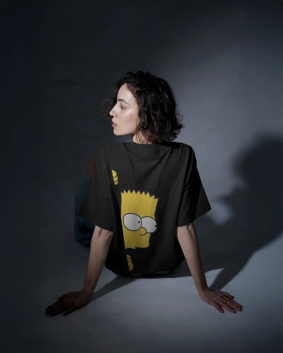 Back view of a female model with short, dark, wavy hair wearing a black baggy t-shirt in a spotlight studio setting. The back of the shirt features a large, bright yellow graphic of Bart Simpson looking over the shoulder of the wearer.