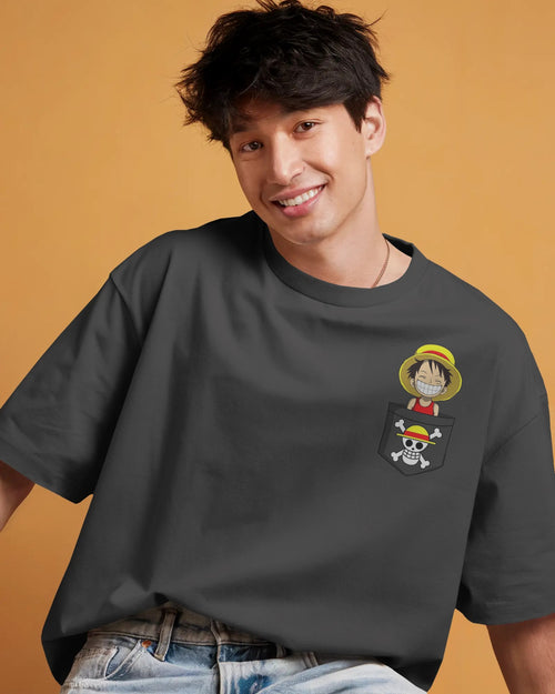 Straw Hat Salute One Piece Brotherhood Oversized T-Shirt - Cheerful male model wearing a dark gray oversized t-shirt with a small graphic on the chest pocket area. The graphic features a stacked design of Luffy's straw hat and the Jolly Roger skull. The background is a warm orange color.
