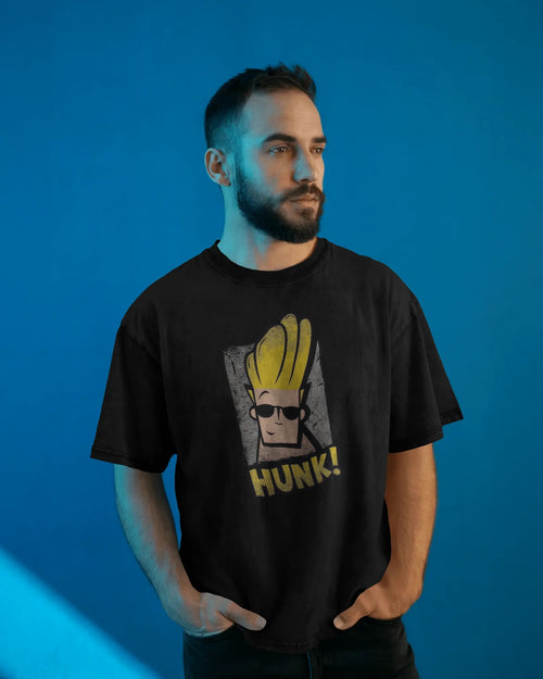 Front view of a bearded man wearing a black, acid-wash, oversized t-shirt with a Johnny Bravo graphic featuring the word "HUNK!" on a vibrant blue background. This mockup displays the relaxed fit and Retro Cartoon Streetwear style of the Johnny Bravo Oversized Tee for Bold Souls.