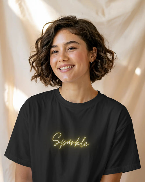Front view of a smiling female model with short, curly brown hair wearing a black t-shirt that features the word "Sparkle" written in a glowing gold script font on the chest. The background is a soft, curtained beige fabric, lit by natural light.