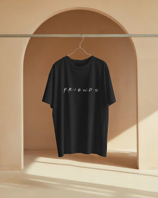 A black relaxed streetwear style tee is shown hanging on a hanger on a metal rod, set within an arched niche against a beige wall with soft sunlight. The front of the shirt displays the classic white "F.R.I.E.N.D.S" logo.