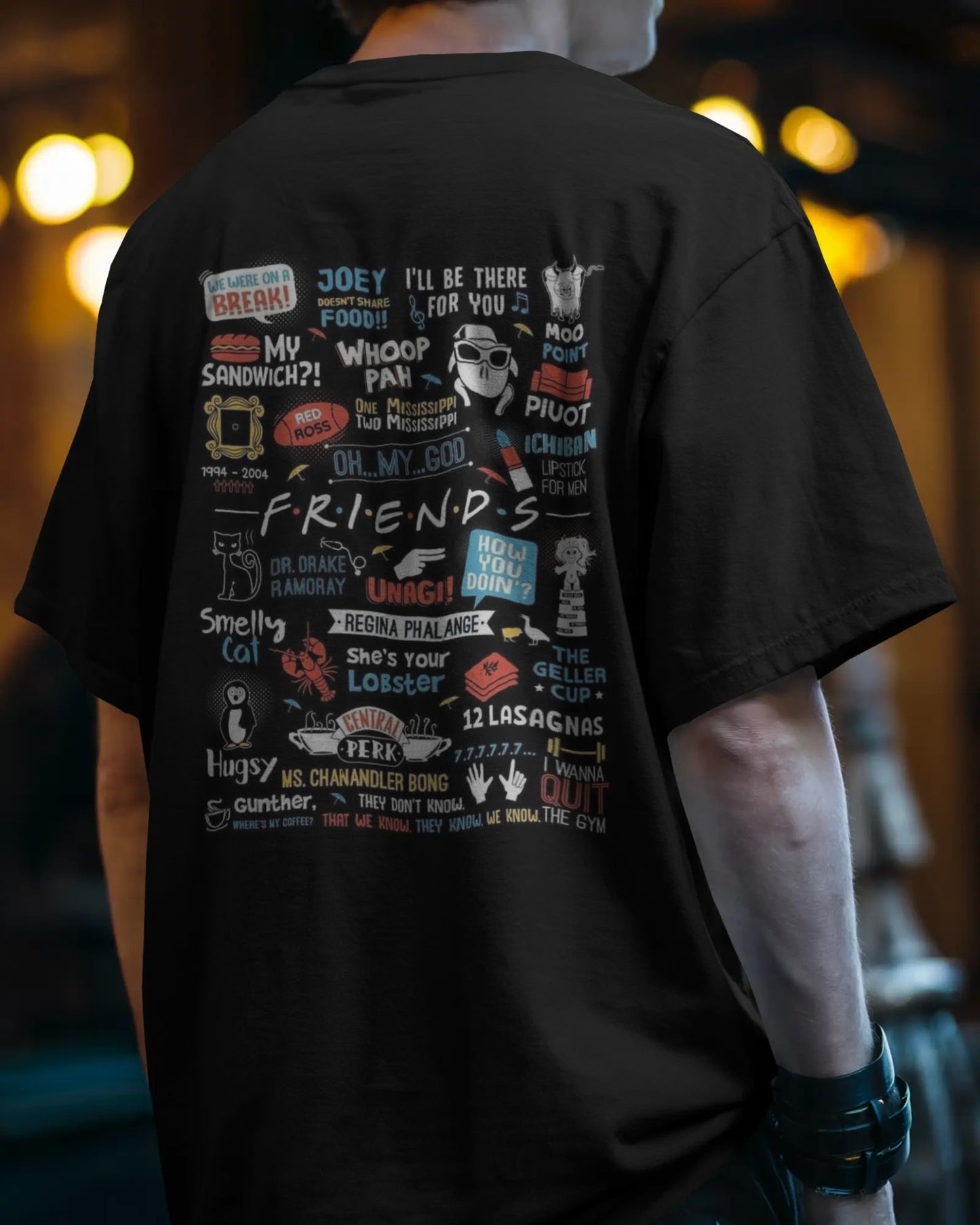 Back view of a male model wearing a black relaxed streetwear style tee with wristbands, in a moody, evening setting with warm bokeh lights. The back of the shirt features a large, colorful collage of iconic F.R.I.E.N.D.S